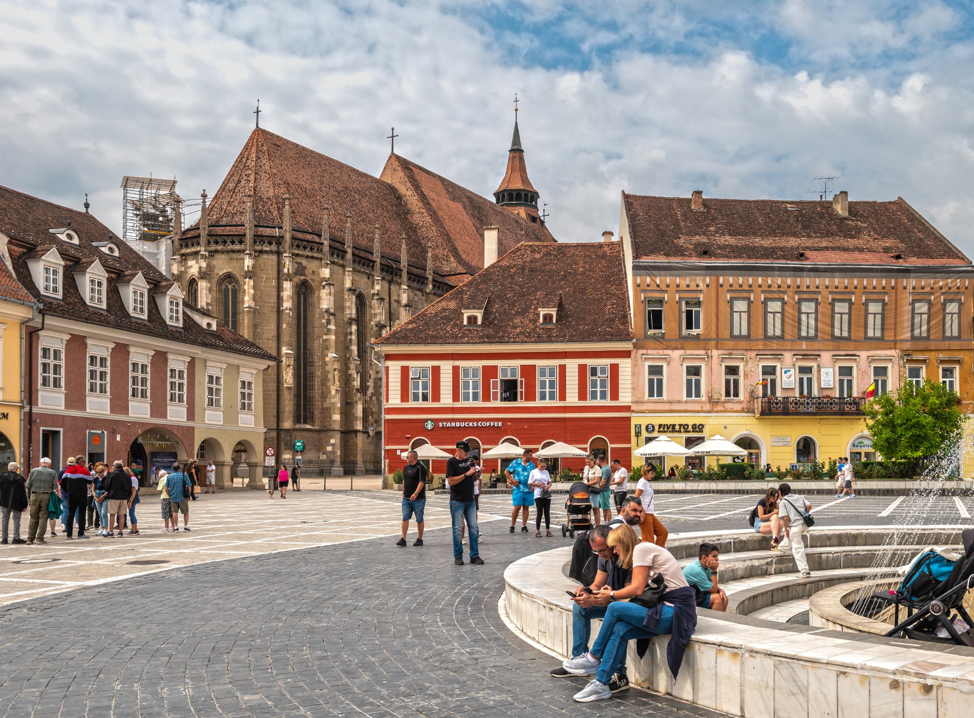 Brașov’daki Konsey Meydanında çeşmenin yanında dinlenen gençler, arkada Kara Kilise
